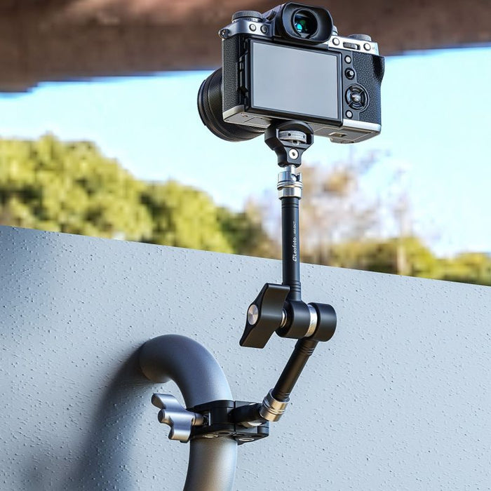 A black handheld gimbal with a mounted camera, shown outdoors attached to a wall under a bridge with trees and sky in the background.