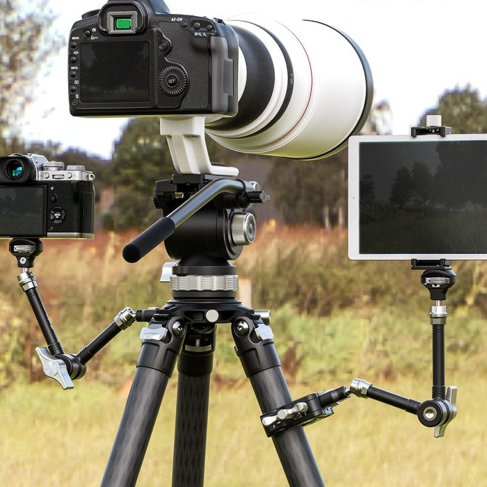 A black camera tripod set up outdoors in a grassy field, holding a large white telephoto lens on top. A tablet is mounted on a small black arm to the right, and another camera is attached on the left.