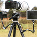 A black camera tripod set up outdoors in a grassy field, holding a large white telephoto lens on top. A tablet is mounted on a small black arm to the right, and another camera is attached on the left.
