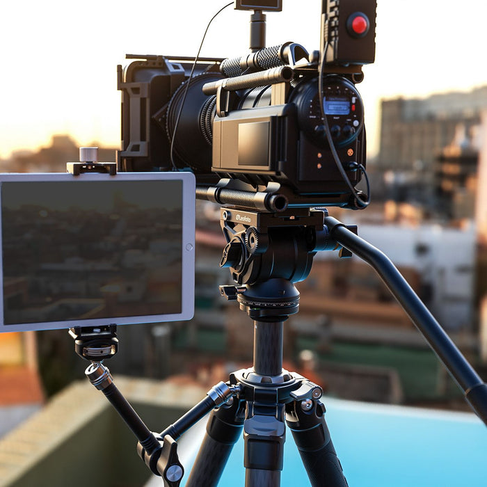 A professional video camera mounted on a tripod with a tablet attached to the side, set against a blurred outdoor cityscape at sunset.