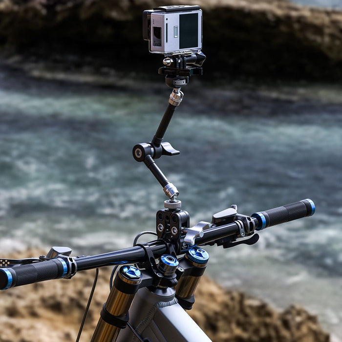 A close-up of a bicycle handlebar setup with a mounted action camera on a flexible mounting arm, photographed outdoors with blurred water and rocks in the background.