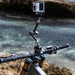 A close-up of a bicycle handlebar setup with a mounted action camera on a flexible mounting arm, photographed outdoors with blurred water and rocks in the background.