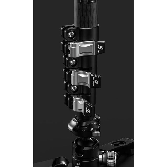 Close-up of a camera tripod head with a carbon fiber pole section and multiple silver and black locking clamps, shown against a dark studio background.