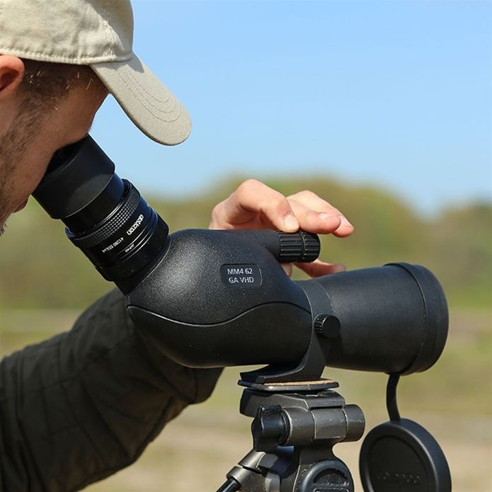 A close-up lifestyle shot of a person using a black spotting scope monocular telescope mounted on a tripod outdoors under a blue sky.