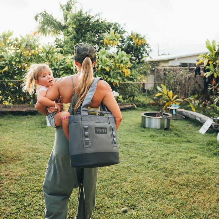 Woman carrying a child while wearing a YETI cooler bag in a backyard setting